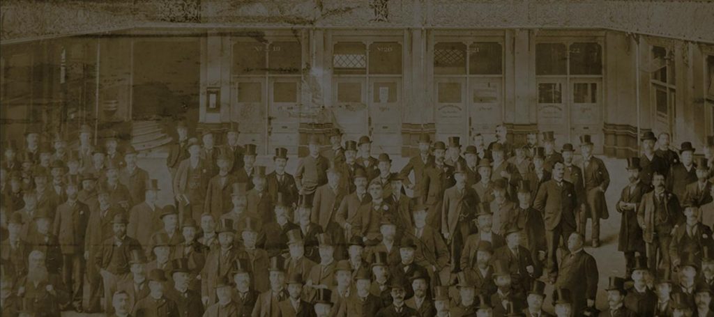 Bennett Opie - Black and white image of a large group of men in suits, ties and hats standing in a factory